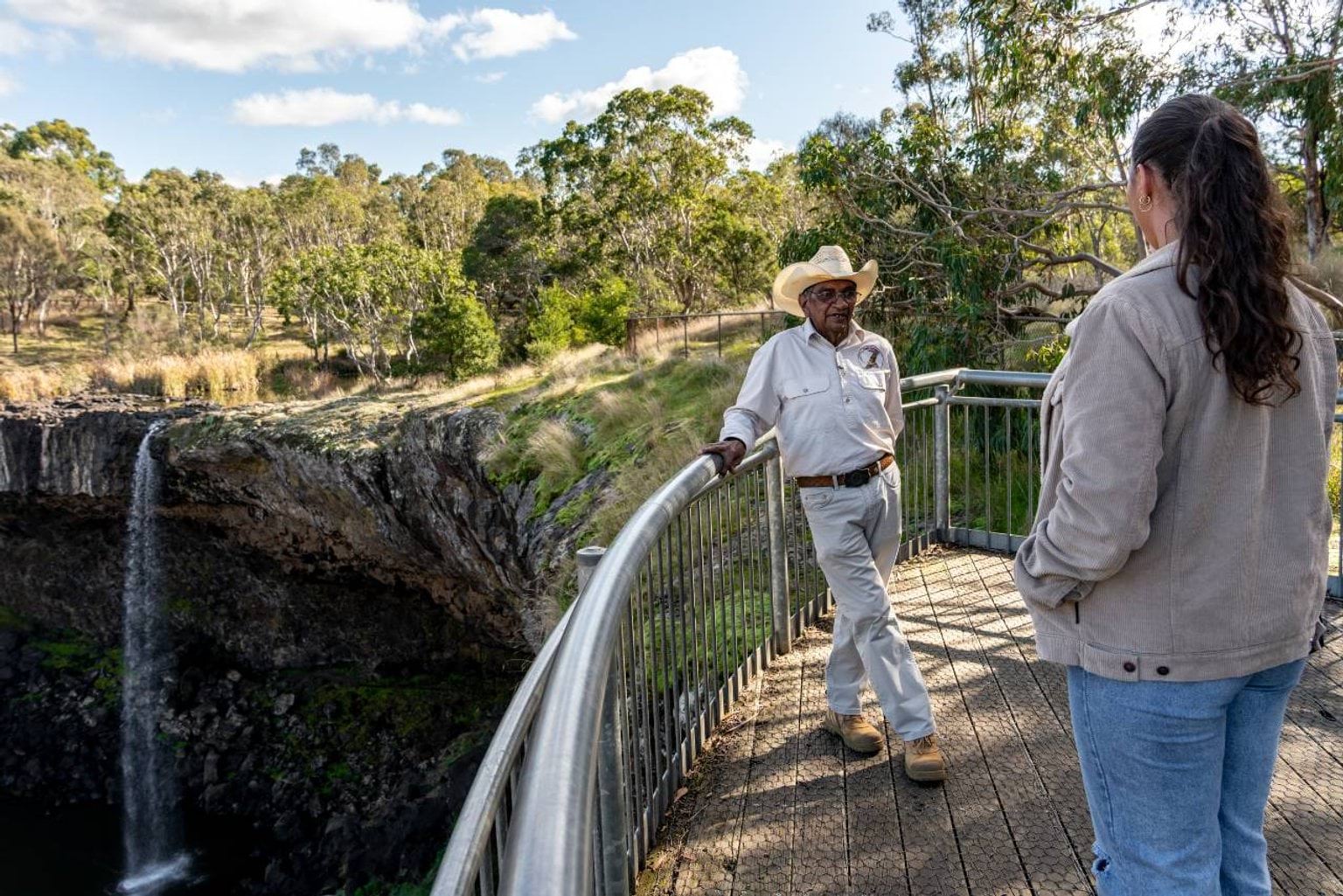 An Aboriginal tour guide wearing a cowboy hat stands on a viewing platform to a waterfall talking to a woman who watches the landscape